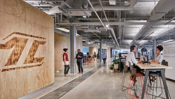 Open-plan office with an exposed-services ceiling, plywood feature wall and colleagues talking by a standing-height bench