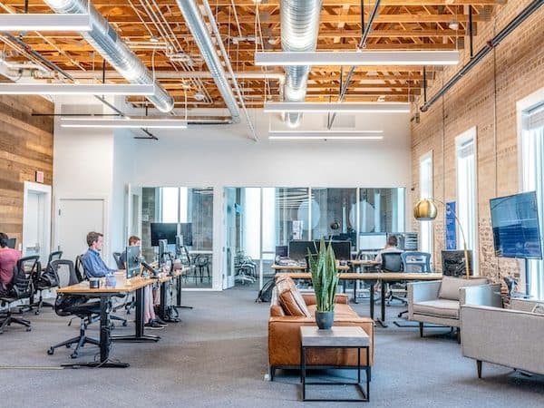 Open-plan office in a converted building with exposed timber deck and brick, bench desks and a tan leather sofa
