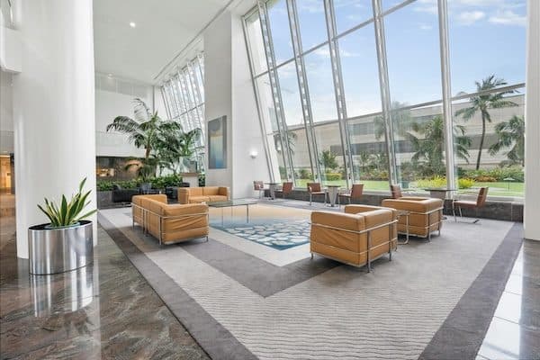 Double-height lobby with a sloped glass wall, tan leather club chairs on a grey rug, palms and polished stone floor