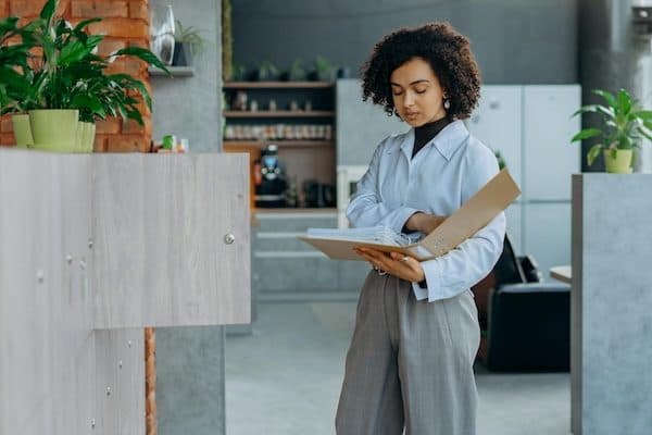 Person holding an open binder beside light-wood storage cabinets in an office kitchen area with plants and brick