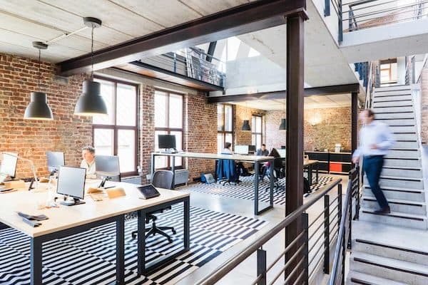 Open-plan office in a converted brick building with bench desks, black pendant lights, a mezzanine and steel stair