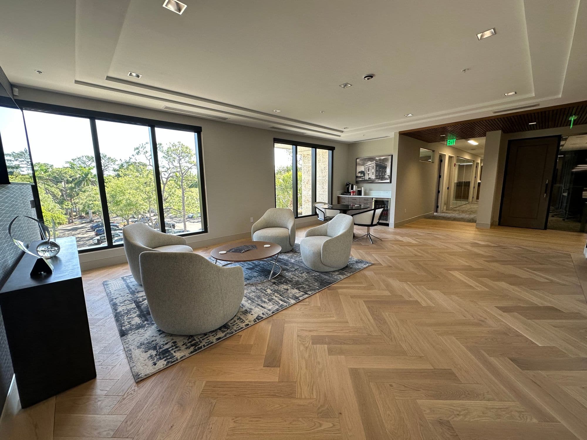 Wider lounge/lobby angle: light-gray swivel chairs around coffee table on rug, herringbone floor, large windows on left, framed art and hallway extending right with green exit sign visible.