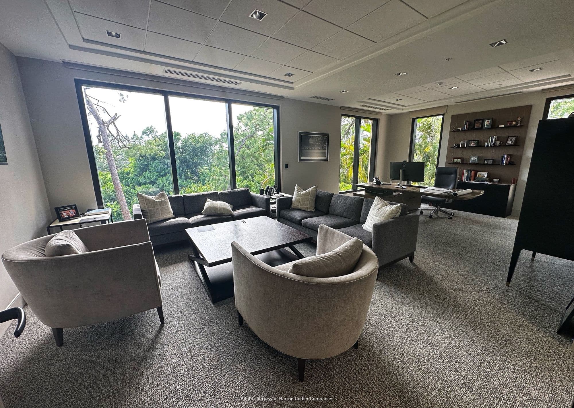 Executive office: gray sectional sofa and three light-gray swivel armchairs around dark coffee tables, full-height windows treetop view, executive desk with built-in bookshelves on right.