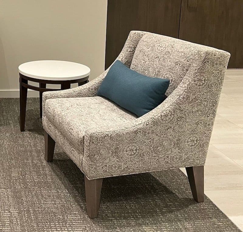 Single gray paisley/damask armchair with small blue lumbar pillow on textured gray carpet, small round white-top side table on dark stem to the left, dark walls behind.
