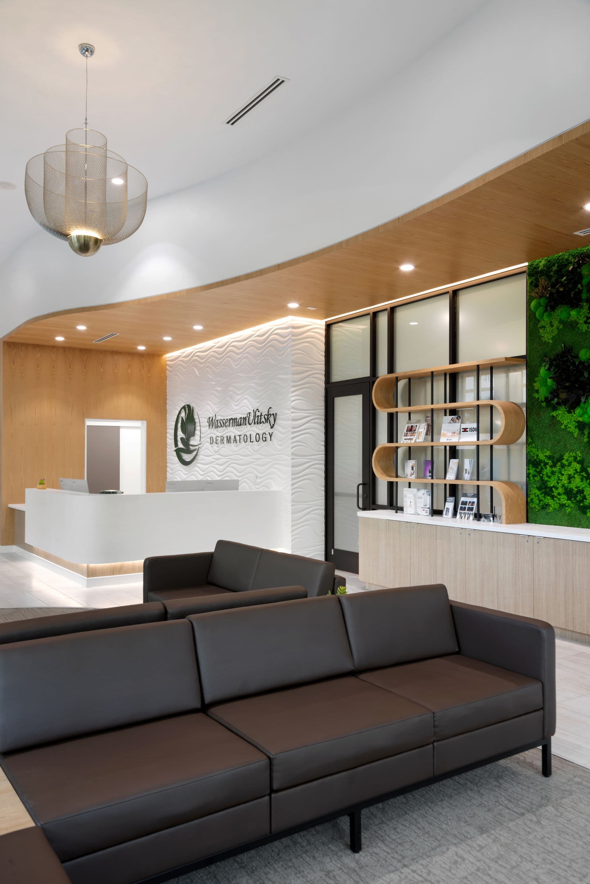 Reception lobby alt angle: dark brown three-seat sofa in foreground, reception desk and curved cove ceiling mid-ground, designer pendant overhead, brand wall and door at far end.