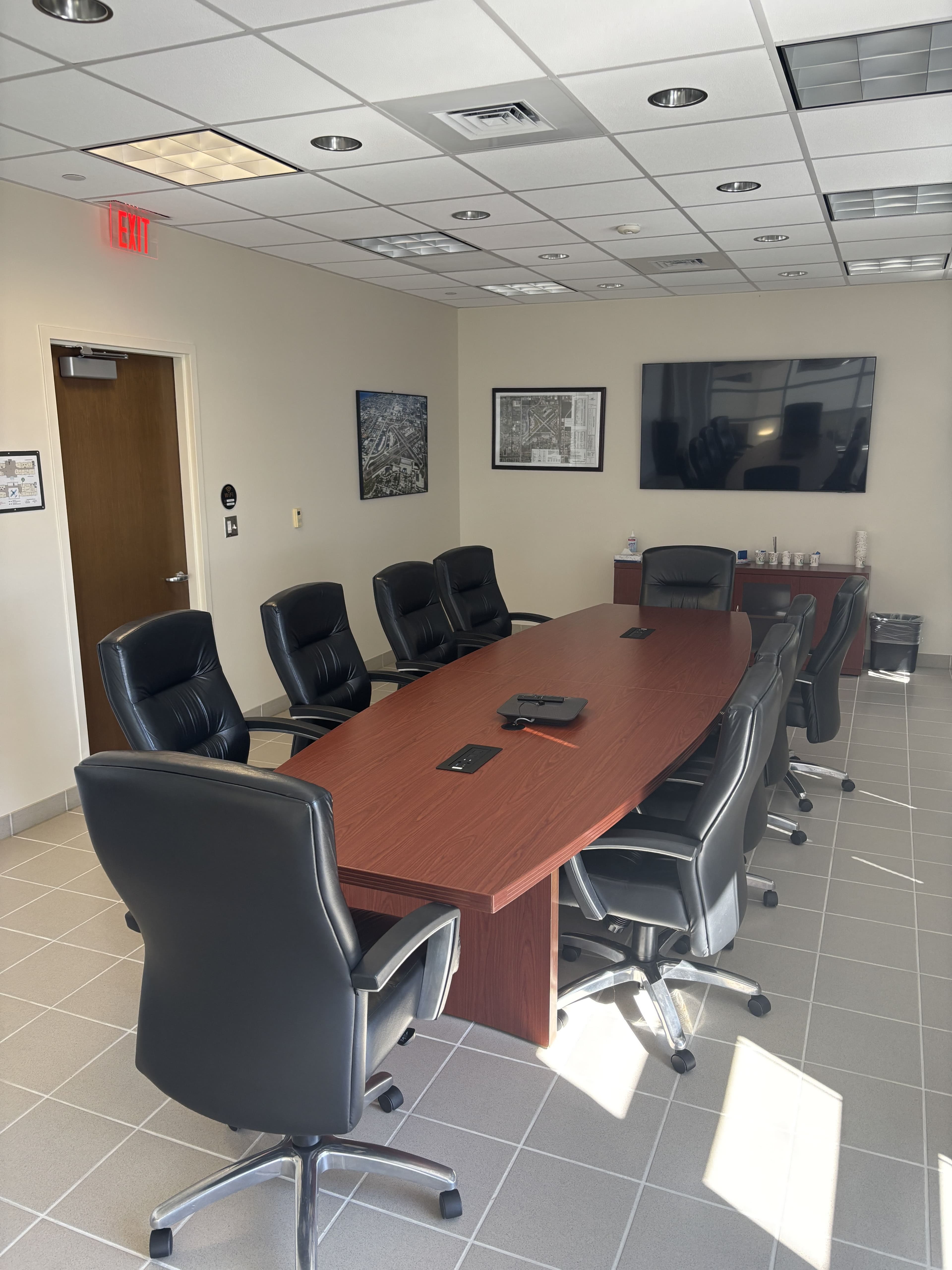 Same conference room, tighter angle: cherry-wood table edge in foreground, 4-5 black executive task chairs around it, framed photos and art on white tile wall, fluorescent ceiling.