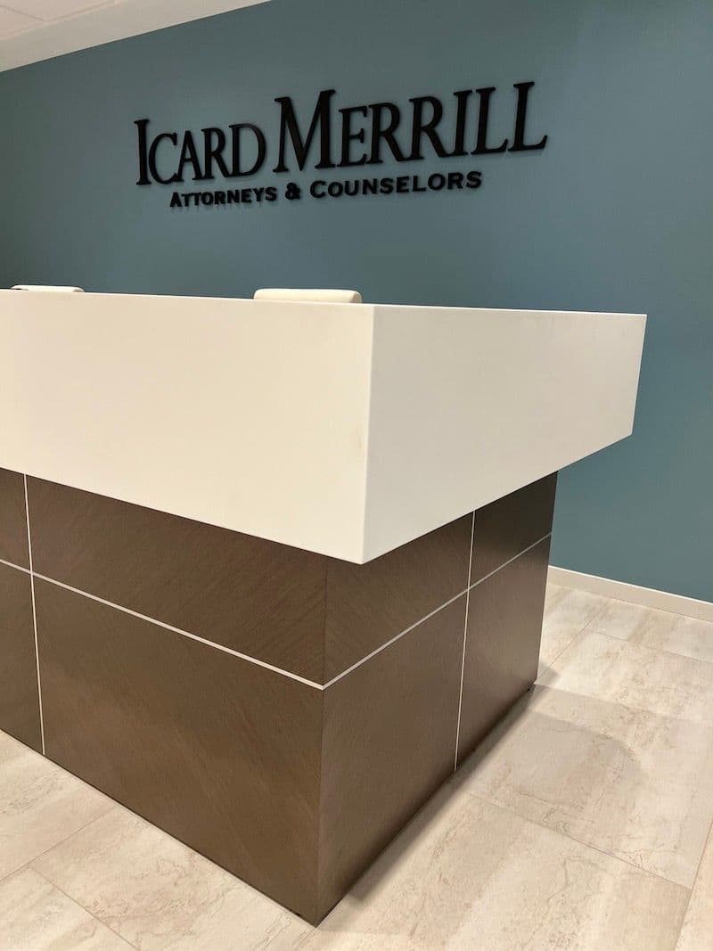 Reception desk corner detail: white quartz cap, dark-wood base with thin reveal line, "Icard Merrill" lettering visible above, travertine floor below.