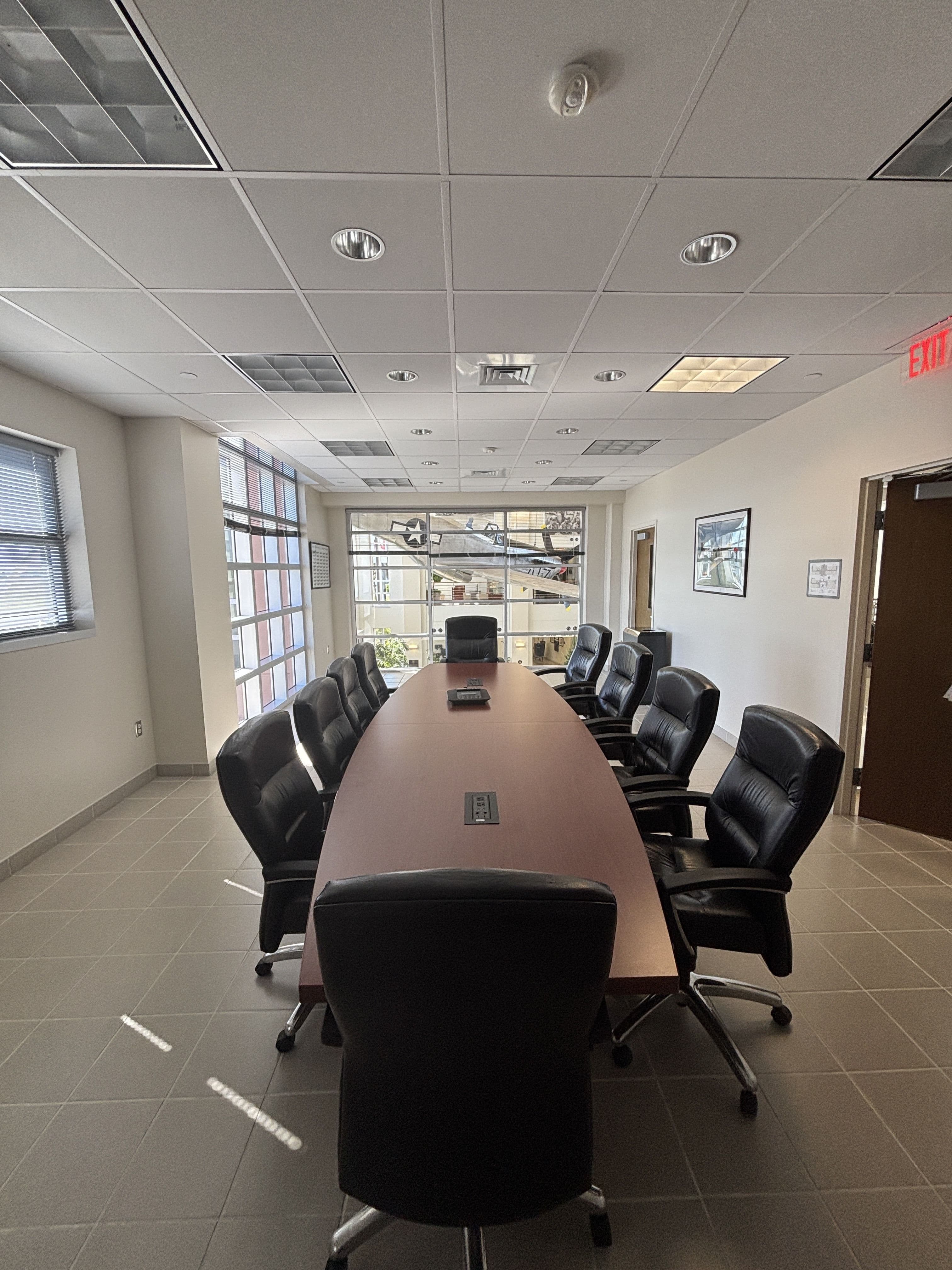 Same conference room, end-of-room angle: full table length visible toward window wall with vertical blinds and view to airfield/jetway, exit sign visible, full set of black executive chairs.
